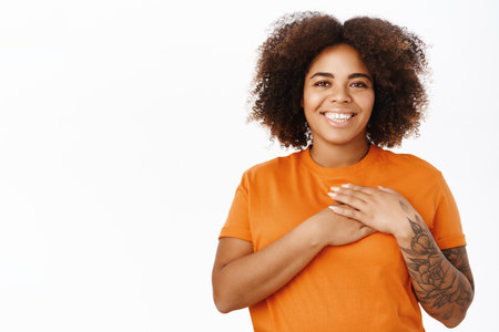 With all her heart. Dreamy smiling african american woman holds hands on chest with caring face expression, standing over white backgroundの写真素材