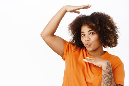 Portrait of cute and silly african american woman frames her head inside arms, pucker lips kissing, standing in orange tshirt over white backgroundの写真素材