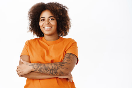 Portrait of confident smiling young african american woman, cross arms, she has tattoos on her arm, looks happy, stands in orange tshirt over white backgroundの写真素材