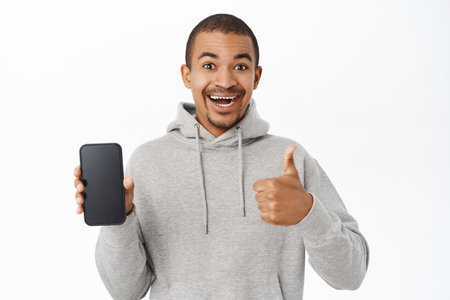 Enthusiastic arab guy shows mobile phone screen and thumbs up, recommends website, telephone application, stands over white backgroundの写真素材