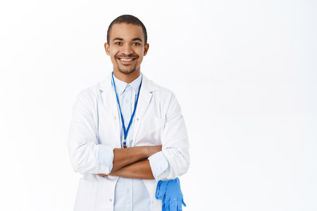 Handsome arab doctor in white coat, cross arms on chest, looks professional, stands over white backgroundの写真素材