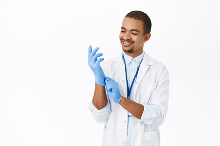 Smiling happy doctor put on medical gloves for patient checkup in hospital, standing over white backgroundの写真素材