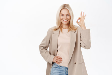 Happy smiling older woman, 50 years old, mature female model, shows okay, ok sign in approval, recommending smth, standing over white backgroundの写真素材
