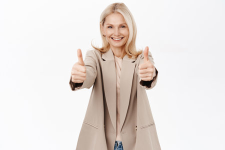 Smiling blond mature woman, older lady shows thumbs up, like and approve, recommends smth, stands over white backgroundの写真素材