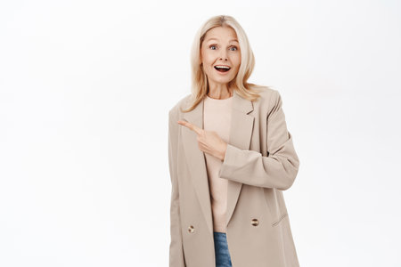 Surprised middle aged woman, blond lady 50 years old, pointing finger left, looking amazed, standing over white backgroundの写真素材