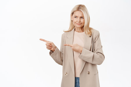 Skeptical middle aged woman with blond hair, pointing and looking left with disappointed face, standing over white backgroundの写真素材