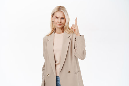Disappointed woman in elegant trench coat, frowning upset, pointing finger up on advertisement, standing over white backgroundの写真素材