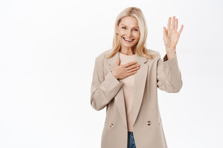 Smiling mature blond woman, saying hello, introduce herself, hi gesture with friendly happy face, standing over white backgroundの写真素材