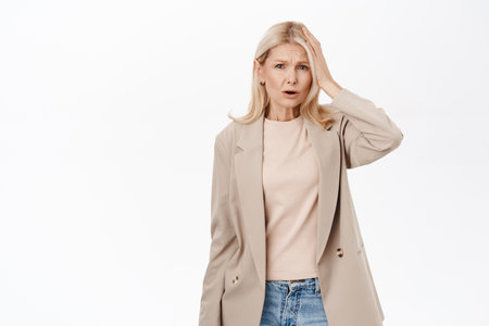 Portrait of mature lady looks confused, gasping and looking worried, forgot smth, stands in trench coat over white backgroundの写真素材