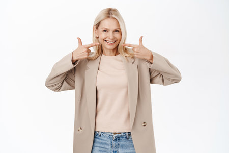 Beautiful and healthy mature lady, elegant senior woman pointing fingers at her white perfect smile, standing over white backgroundの写真素材