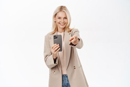 Elegant, beautiful middle aged woman recording video on smartphone, taking photos on mobile phone camera, smiling, standing over white backgroundの写真素材