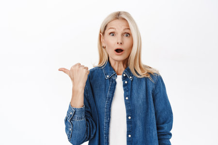 Close up portrait of surprised elegant senior woman, 50s years, pointing finger left, showing advertisement, promo offer, standing over white backgroundの写真素材