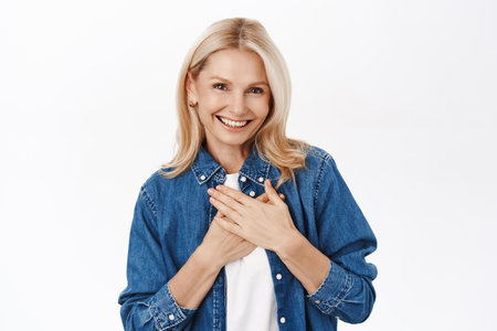 Portrait of smiling blond senior woman, grandmother looking with love and care, holding hands on heart, standing over white backgroundの写真素材