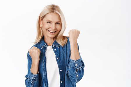 Yes, success. Excited blond mature woman celebrating, makes fist pump and smiles satisfied, achieve goal, triumphs, stands over white backgroundの写真素材