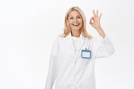 Excellent. Smiling beautiful woman doctor, senior lady in medical uniform, shows okay, ok sign, approves smth, agrees, stands over white backgroundの写真素材