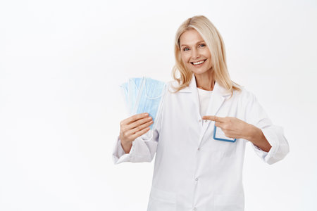 Covid-19 protection. Smiling senior woman doctor, shows medial face masks, recommends wearing personal protective equipment, standing over white backgroundの写真素材