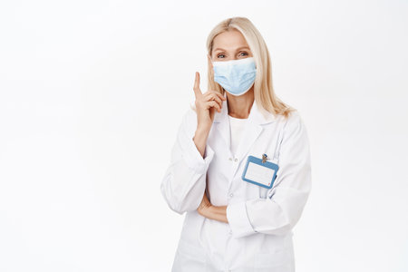 Portrait of smiling healthcare worker, lab employee in medical face mask, points finger up, shows advertisement, stands over white backgroundの写真素材