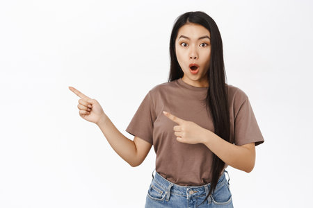 Portrait of amazed asian girl pointing fingers left, showing awesome promo offer, best deal, standing over white backgroundの写真素材