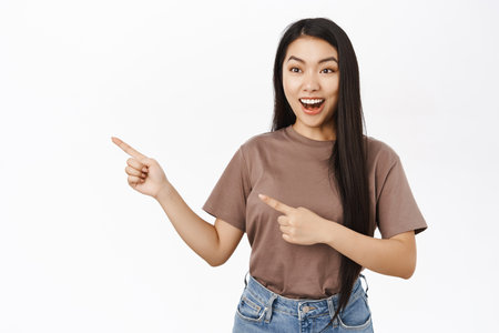 Portrait of amazed asian girl pointing fingers left, showing awesome promo offer, best deal, standing over white backgroundの写真素材