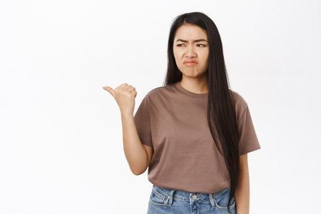 Jealous asian girl, disappointed woman pointing finger left, grimacing displeased, standing over white backgroundの写真素材