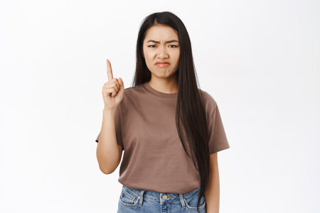 Jealous asian girl points up, grimaces from regret, disappointment, stands over white studio backgroundの写真素材