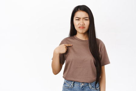 Sad asian woman pointing at herself, sulking and grimacing upset, being unconfident, standing over white backgroundの写真素材