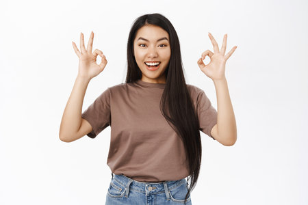 Portrait of cute asian girl shows okay, ok, excellent gesture, smiling pleased, praising and complimenting, say yes, white backgroundの写真素材
