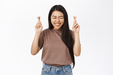 Portrait of hopeful asian girl, prays, makes wish and stands with crossed fingers, anticipates, white studio backgroundの写真素材