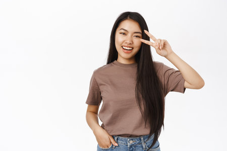 Portrait of smiling asian woman shows peace, v-sign and looks happy, shows positivity and joy, stands in tshirt over white backgroundの写真素材