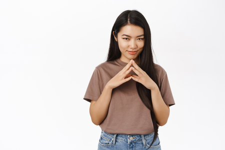 Image of smiling coy asian girl, planning, scheming, steeple fingers with thinking face, white studio backgroundの写真素材
