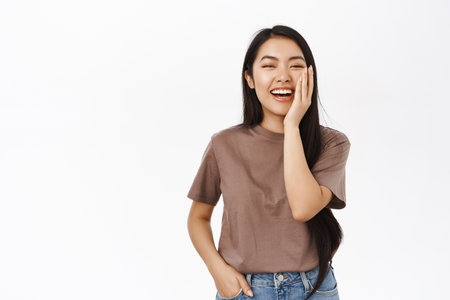 Portrait of beautiful laughing asian girl looking positive, happy face expression, relaxed pose, standing in brown tshirt over white backgroundの写真素材