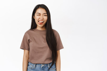 Cool young asian girl shows tongue, winks and express joy, carefree emotion, stands in tshirt over white backgroundの写真素材