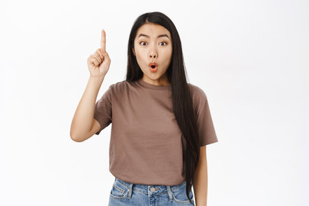 Girl pitching an idea. Excited asian woman raising finger up, saying suggestion, has plan, standing over white backgroundの写真素材