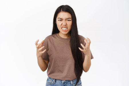 Angry asian teen girl shakes hands, grimacing outraged, furious, express rage and anger, stands over white backgroundの写真素材