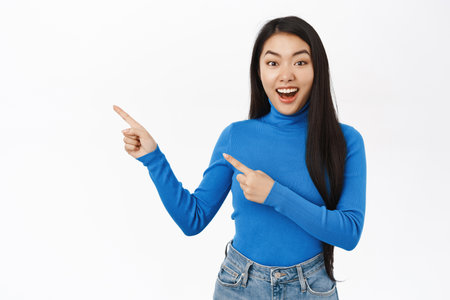 Portrait of smiling asian woman pointing fingers left, showing amazing news, promo banner, offer, standing over white studio backgroundの写真素材
