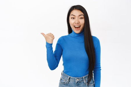 Portrait of smiling asian woman pointing fingers left, showing amazing news, promo banner, offer, standing over white studio backgroundの写真素材
