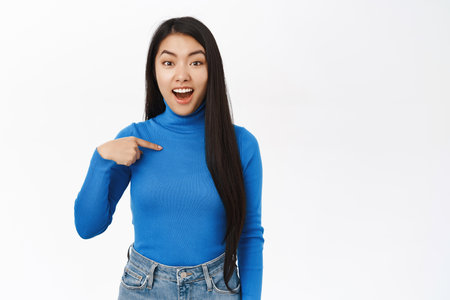 Excited korean woman pointing at herself with surprised, amazed face, express surprise and disbelief, being chosen, stands over white backgroundの写真素材