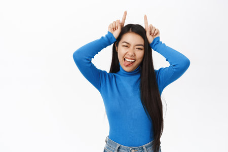 Enthusiastic asian woman shows devil horns, fingers on head, smiles sassy and extends tongue, playful and flirty emotion, white backgroundの写真素材