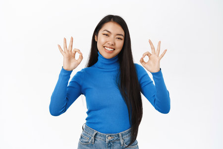 Smiling asian girl shows okay, ok sign in approval, praise excellent promo deal, recommends company, stands over white backgroundの写真素材