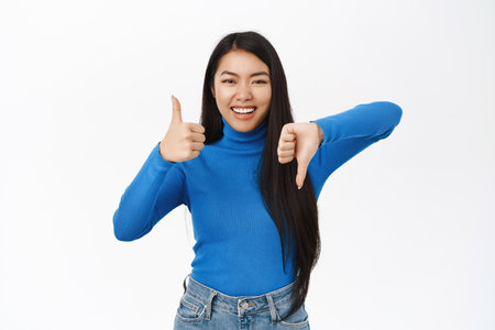 Like or dislike. Smiling asian girl shows thumbs up and thumbs down, makes choice, rate and give feedback, makes decision, stands over white backgroundの写真素材