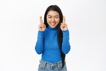 Nervous asian girl praying, wishing, waiting with concerned, worried face, cross fingers for good luck, white backgroundの写真素材