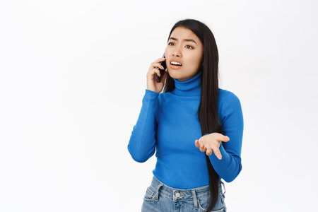 Portrait of korean woman with confused face talking on mobile phone, having a call, talking with puzzled face on telephone, white backgroundの写真素材