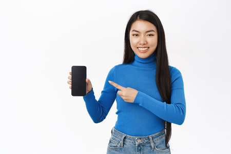 Portrait of beautiful smiling girl pointing finger at mobile phone, showing application on smartphone, standing over white backgroundの写真素材