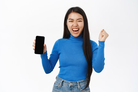 Enthusiastic korean woman shows mobile phone screen, looks excited and surprised, winning on smartphone, standing over white backgroundの写真素材