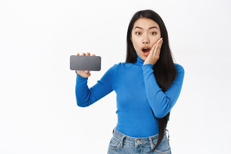 Technology. Smiling asian woman demonstrates smartphone app screen, shows horizontal mobile phone interface, application, stands over white backgroundの写真素材