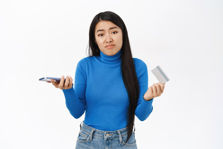 Confused asian girl holds smartphone with credit card, shrugs shoulder with puzzled face, white backgroundの写真素材