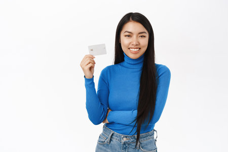Smiling asian woman recommends bank, shows credit card, stands over white studio backgroundの写真素材