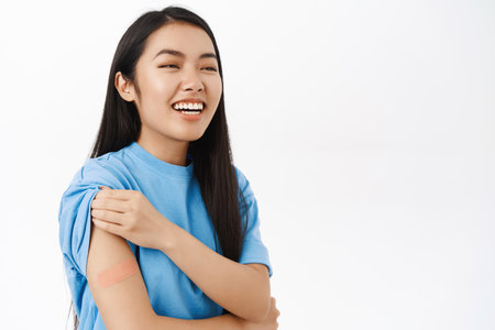 Covid-19 vaccination. Smiling asian healthy girl, has bandaid on shoulder after coronavirus vaccine, standing over white backgroundの写真素材