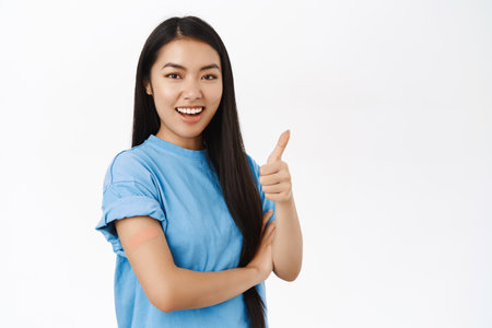 Enthusiastic asian girl got vaccinated, has bandaid patch on shoulder after covid vaccine, smiling and pointing at copy space banner, showing coronavirus info announcementの写真素材