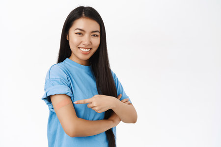 Smiling chinese girl points at shoulder with bandaid, promotes vaccination from covid-19, coronavirus vaccine advertisement, white backgroundの写真素材